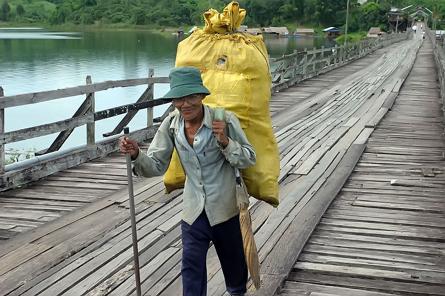 Old man with a big sack of something crossing the wooden footbridge in Sangkhlaburi